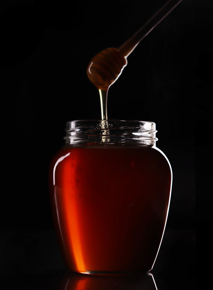 Rich amber honey dripping from a wooden dipper into a glass jar against a dark background.