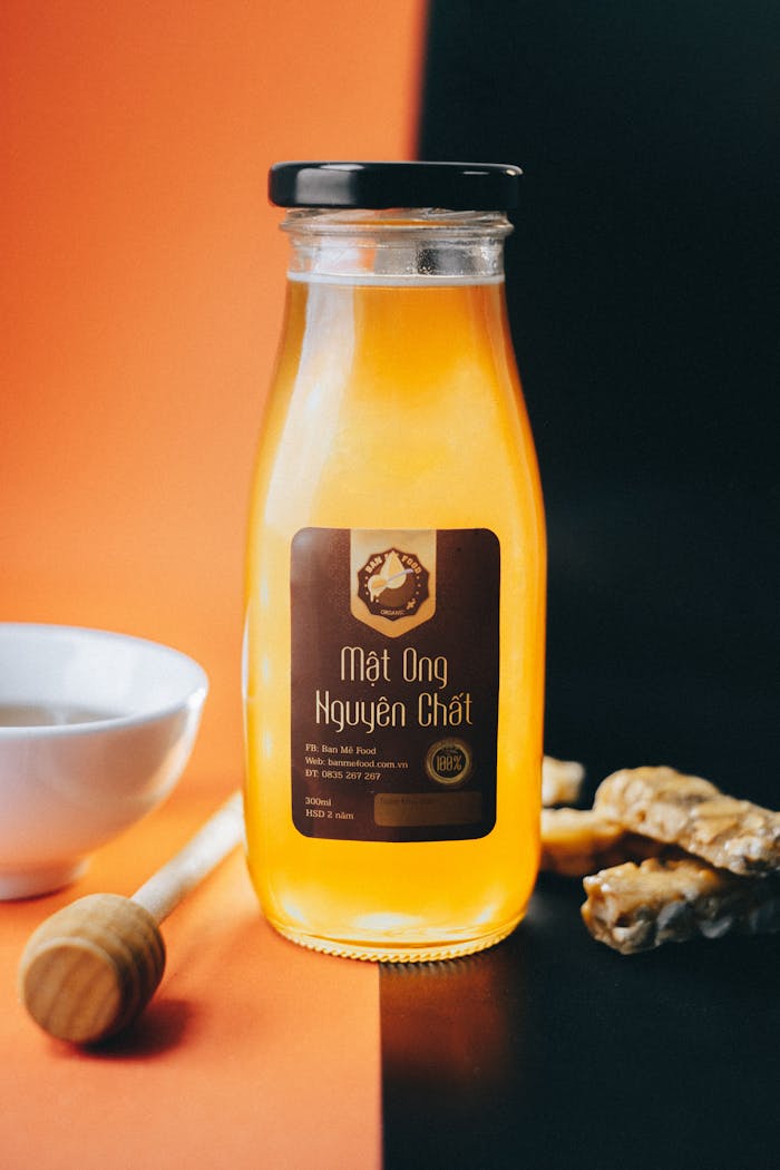 Glass bottle of pure honey with label, placed against two-toned backdrop. Product shot with honey dipper and bowl.