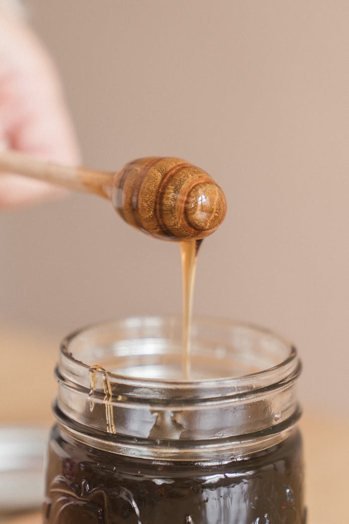 A close-up of honey dripping from a wooden dipper into a glass jar, showcasing its rich texture.